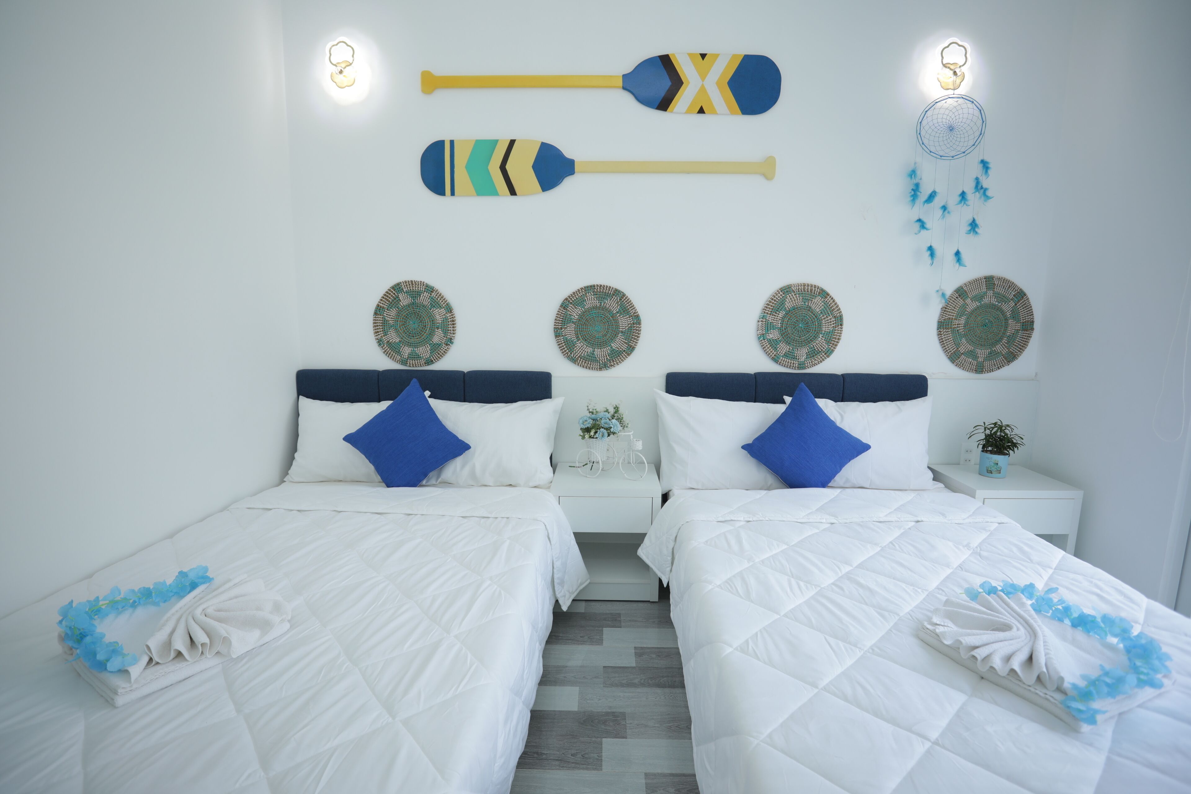 Habitación de hotel con dos camas, ropa de cama blanca, almohadas azules, decoración en la pared y una mesita de noche con una planta.