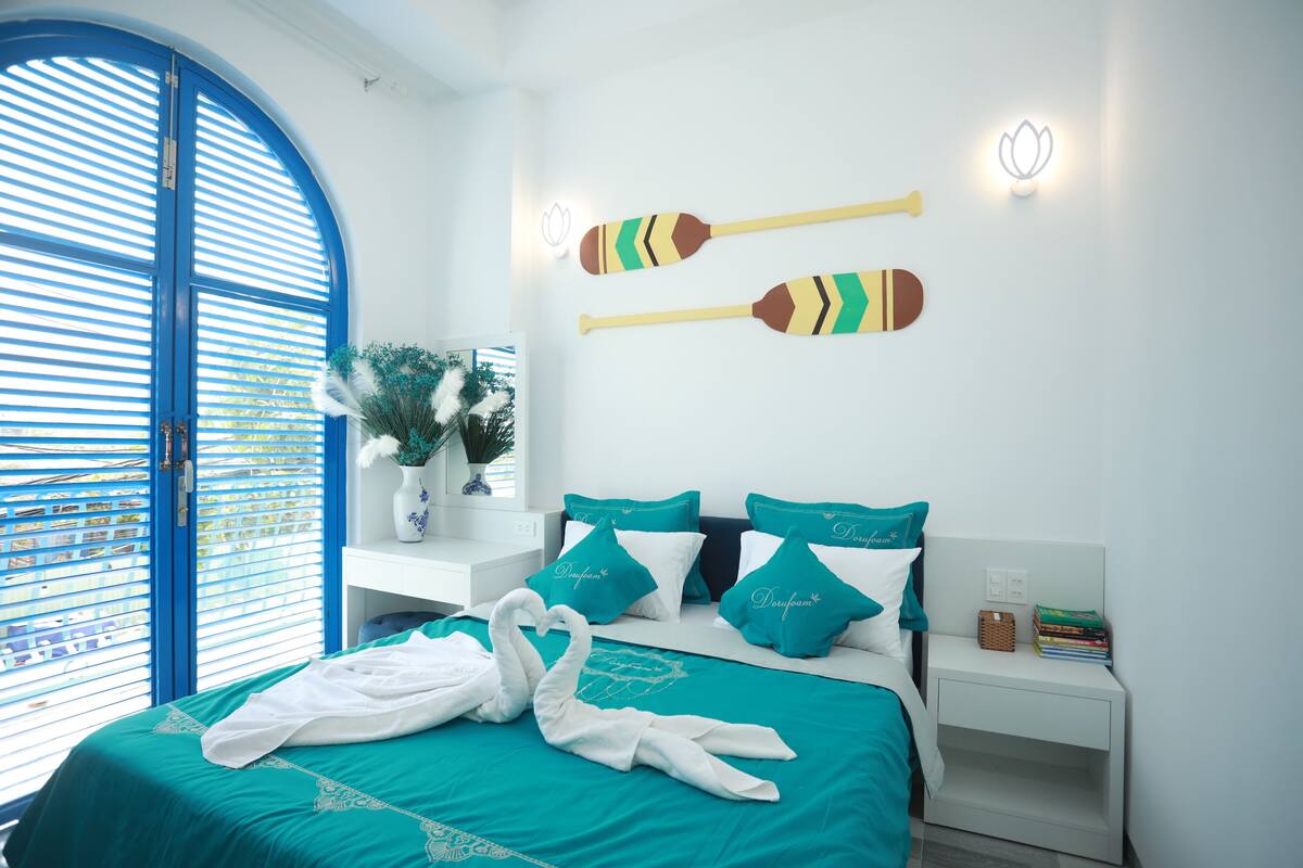 A hotel room with a bed, pillows, a nightstand, a wall-mounted light, and a window with blinds.