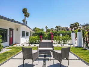 A patio with wicker furniture, a white building with a red door, and a garden with palm trees and pink flowers.