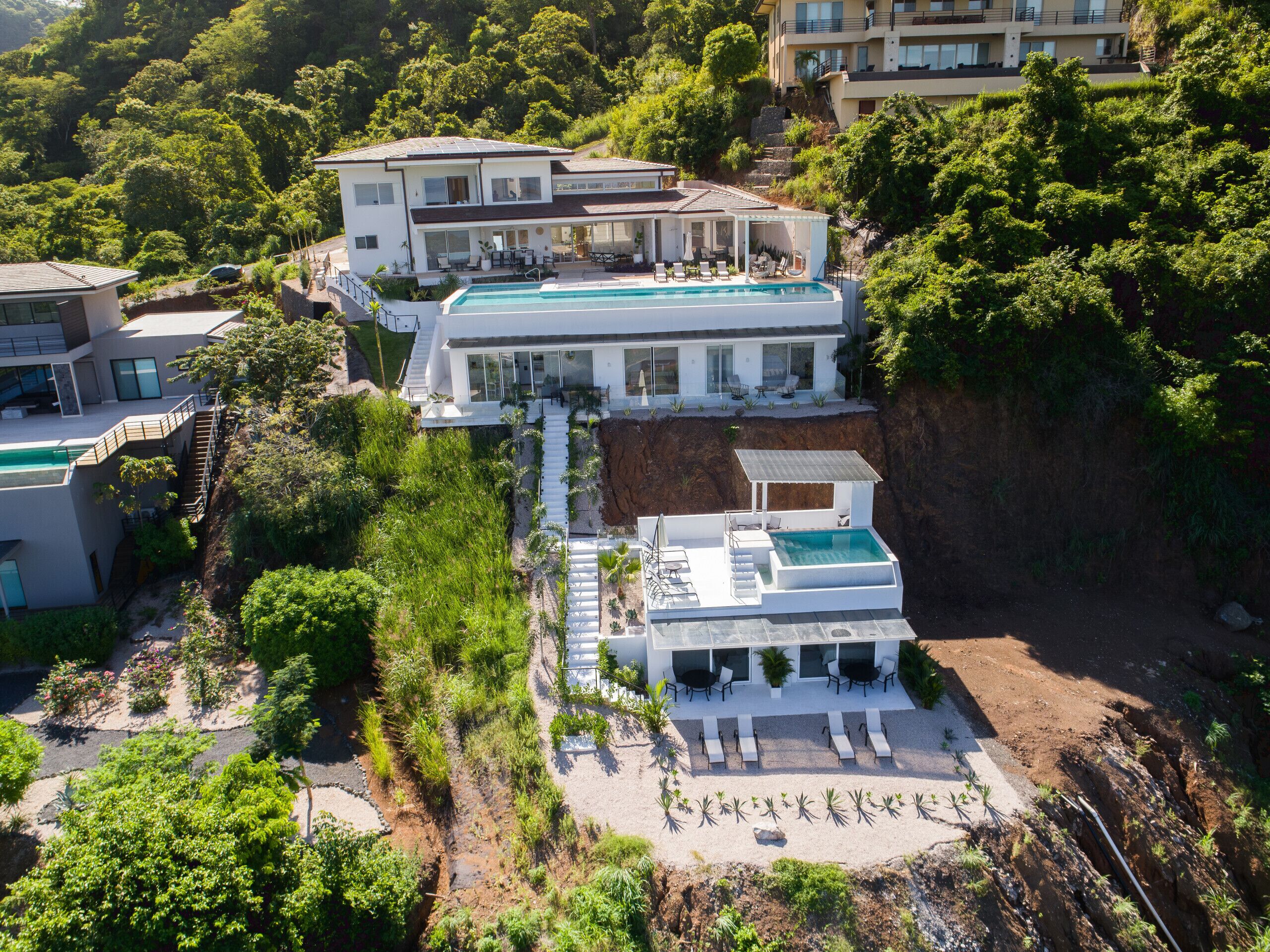 A modern two-story house with a swimming pool, surrounded by lush greenery and a steep hillside.