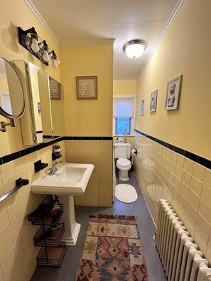 Ein schmales Badezimmer mit Waschbecken, Toilette und Spiegel. An den Wänden befinden sich Bilder in Rahmen und auf dem Boden ein Teppich.