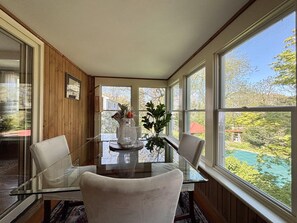 Ein Essbereich mit einem Glastisch, zwei Stühlen, einer Blumenvase und großen Fenstern mit Blick auf Bäume und ein Haus.