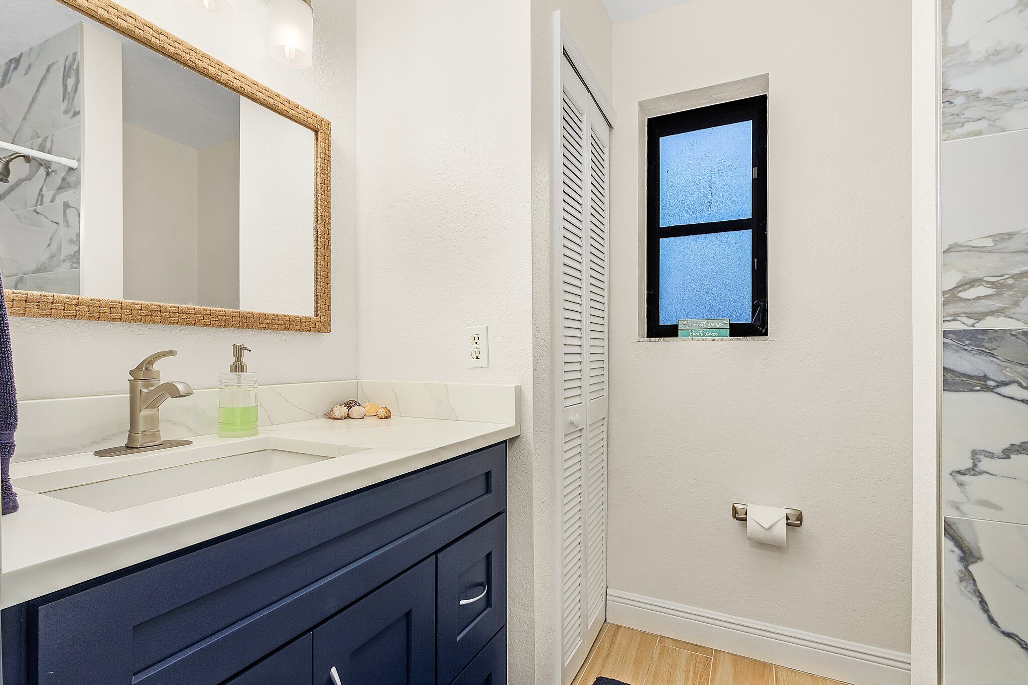 A bathroom with a white sink, dark blue vanity, a mirror with a wicker frame, a window, and a towel rack.
