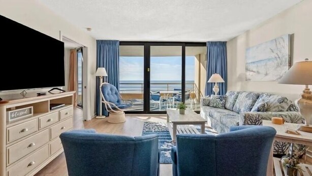 Sala de estar moderna com uma televisão grande, um sofá, uma poltrona e uma varanda com vista para o mar.