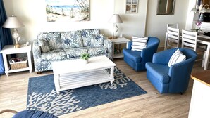 Uma sala de estar com um sofá estampado, uma mesa de centro, duas poltronas azuis, uma mesinha de cabeceira com abajur e um tapete azul.