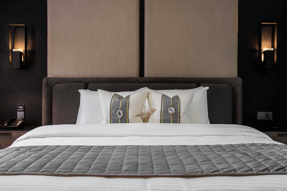 A neatly made bed with a quilted grey bedspread, two pillows with a decorative pattern, and a headboard with a light fixture on either side.
