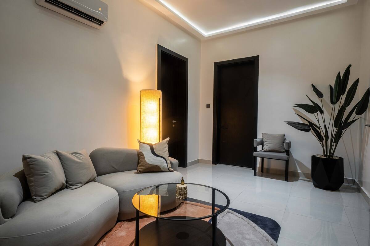 A modern living room with a grey sofa, a glass coffee table, a floor lamp, a potted plant, and a door in the background.