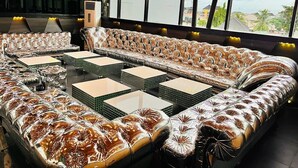 A modern lounge area with a large, patterned sofa and multiple small tables, large windows with a view of buildings and trees, and a contemporary air conditioning unit. - Dluxe Premium Hotel (Port Harcourt)