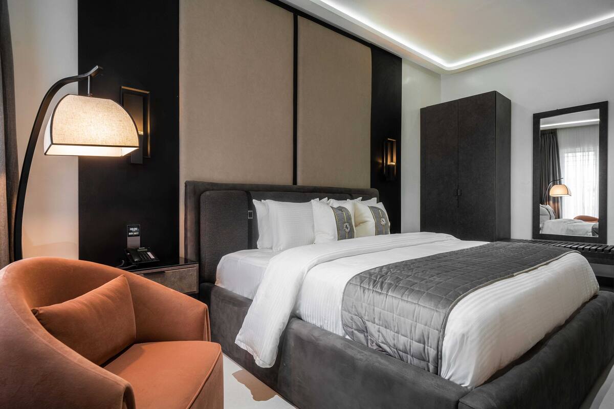 A modern hotel room with a large bed, a desk with a lamp, a chair, and a wardrobe.