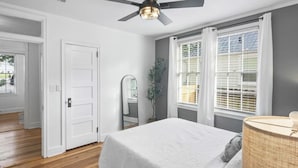 Un dormitorio con cama, ventilador de techo, ventana con persianas y puerta que da a otra habitación.