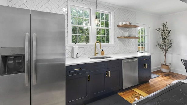 Una cocina moderna con un refrigerador de acero inoxidable, armarios negros con detalles dorados, una encimera blanca y una ventana con vista a follaje.