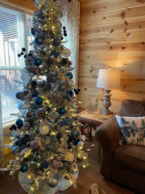 Uma árvore de Natal decorada com enfeites e luzes, um abajur, e um sofá com almofada em um quarto com paredes de madeira.