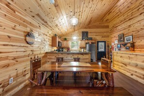 Interior de uma cabana de madeira com uma mesa de jantar central, área de cozinha e um canto de estar acolhedor.