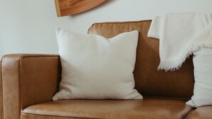 A brown leather sofa with white cushions and a white throw blanket, a wooden paddle leaning against the wall, and a glass coffee table with a woven mat. - Beautiful & Bright Townhome (Westerville)
