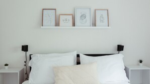 A minimalist bedroom with a white bed, two bedside tables, a nightstand, and a wall-mounted shelf with framed artwork. - Beautiful & Bright Townhome (Westerville)