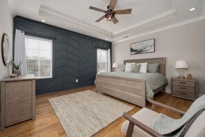 A bedroom with a bed, bedside tables, a desk, a chair, a ceiling fan, and a large window with blinds.