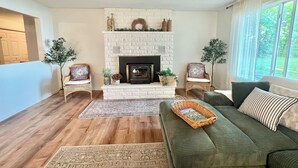 Una sala de estar con chimenea, dos sillones de mimbre, un sofá verde y un ventanal grande.