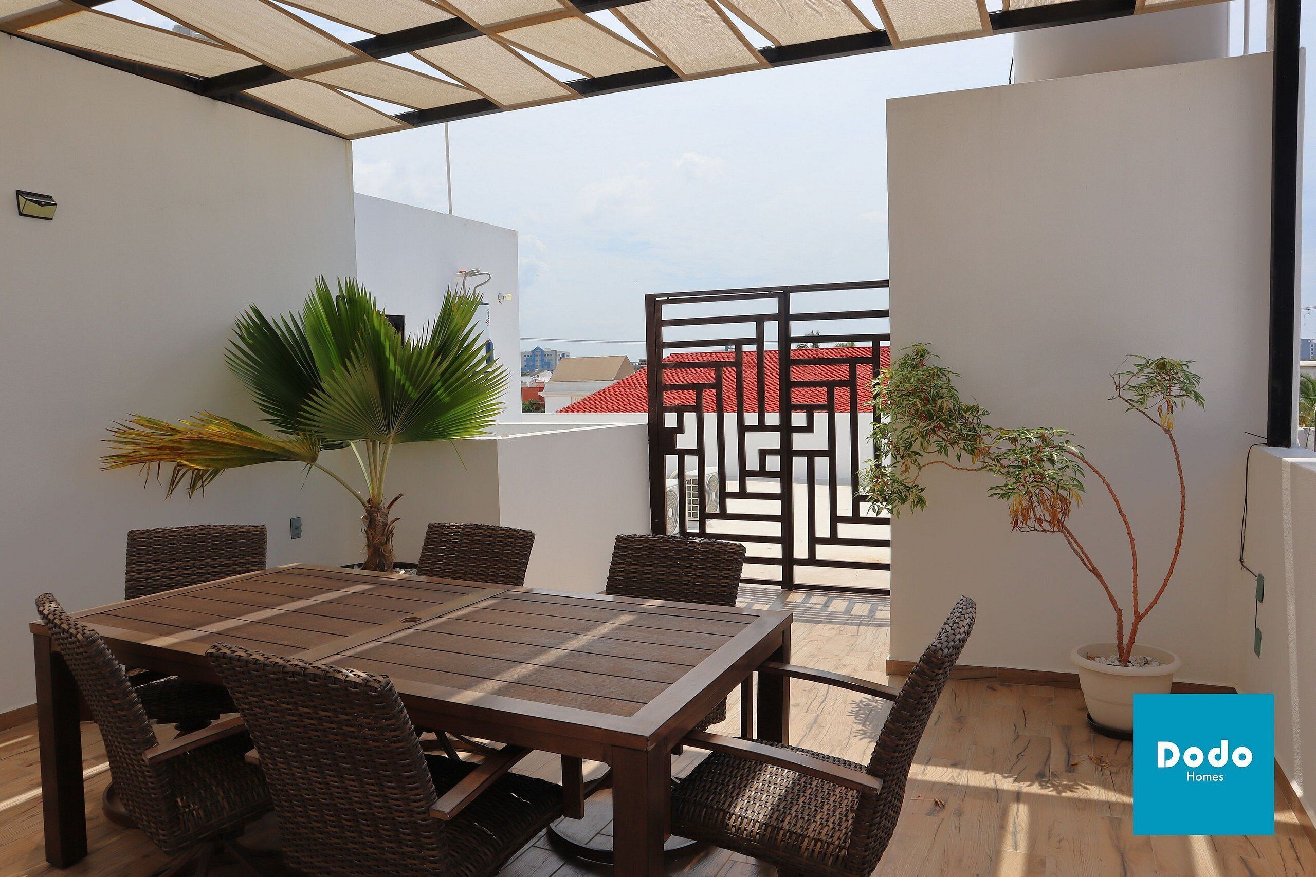 A covered patio with a dining table, wicker chairs, potted plants, and a view of buildings in the background.