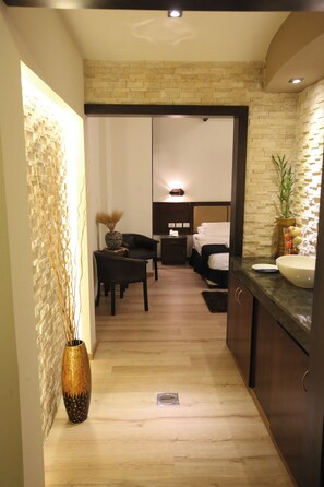 A modern hotel room with a wooden floor, a bed with white linens, a small desk with a chair, a bedside table with a lamp, and a large bathroom with a sink and mirror. - Shepherd Plaza Hotel (Bethlehem)
