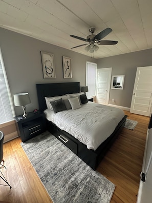 Une chambre à coucher comprenant un lit, des tables de chevet, un ventilateur de plafond, des œuvres d’art, un miroir et une porte.