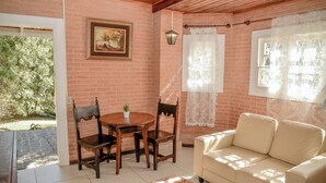 A room with a brick wall, a wooden ceiling, a white sofa, a wooden dining table with chairs, and a window with lace curtains. - Chalé Terrazza  (Campos do Jordão)