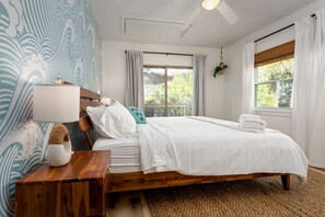 A bedroom with a wooden bed, white bedding, a bedside table with a lamp, and a view of greenery outside.