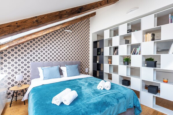 A modern bedroom with a sloped ceiling, a patterned wallpaper, a large bed with a blue cover, and a white shelving unit with various items.