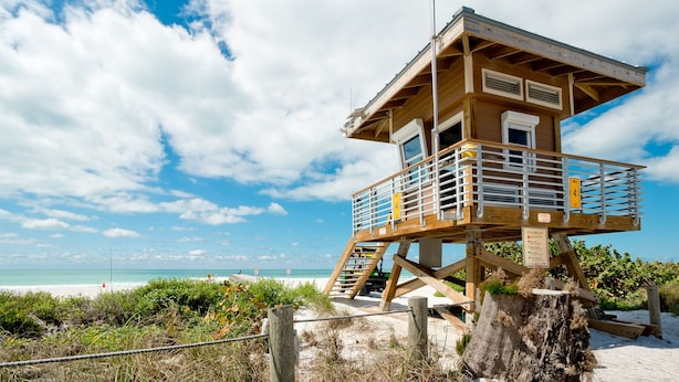 Een uitkijktoren voor strandwachten, met een houten constructie, trappen en leuningen, op een zandstrand met begroeiing op de voorgrond.