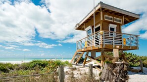 Une tour de surveillance, dotée d’une structure en bois, d’un escalier et de garde-corps, est située sur une plage de sable, avec une végétation au premier plan.