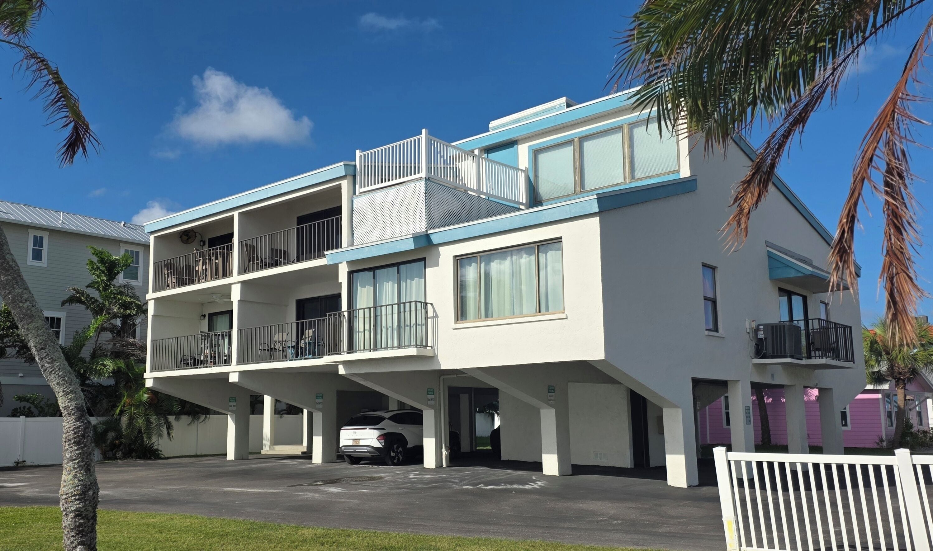 A multi-story residential building with balconies, a parking area, and a white picket fence.