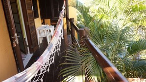A hammock hanging between wooden posts, a white plastic chair, and a table with a blue object on it, all set against a yellow wall and surrounded by lush green foliage. - Recanto Primavera - Camburi (São Sebastião)