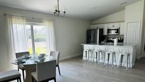 Una cocina moderna con armarios blancos, un refrigerador de acero inoxidable y un horno empotrado. Hay un comedor con una mesa de vidrio y cuatro sillas.