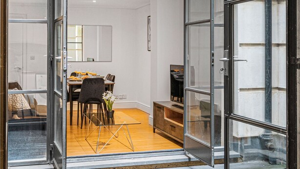 An open glass door leading to a dining area with a table set for two, a chair, and a potted plant.