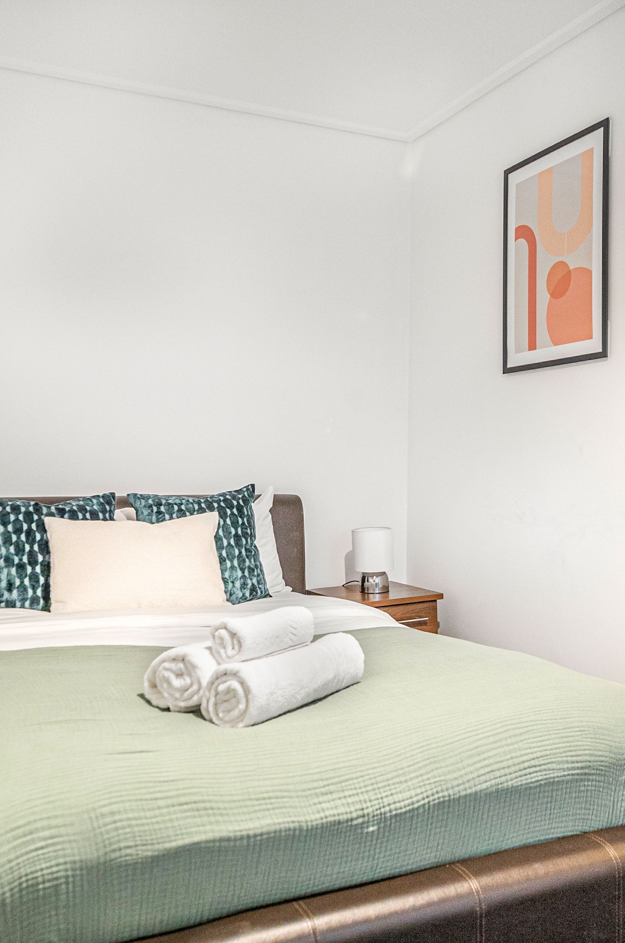 A neatly made bed with a green bedspread and white pillows, a bedside table with a lamp, and a framed abstract artwork on the wall.