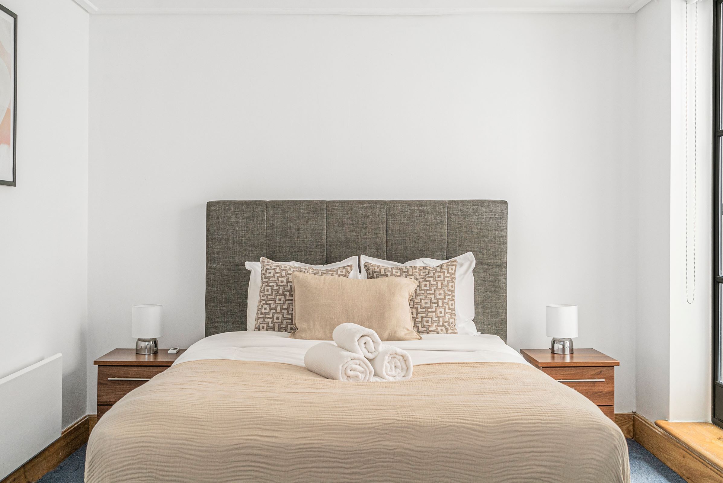 Un lit bien fait, avec une couette beige et des serviettes pliées, une tête de lit rembourrée de couleur grise et deux tables de chevet avec des lampes et un téléphone.