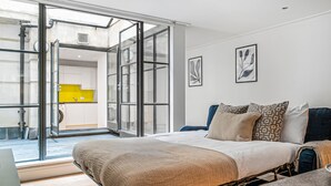 A modern bedroom with a large bed, a desk, and a view of a kitchen through a glass door.