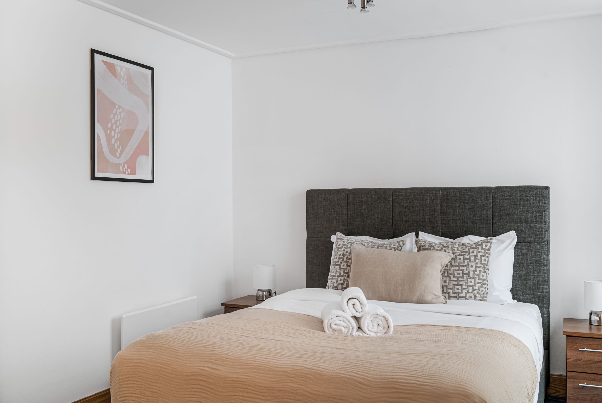 A neatly made bed with a grey headboard, white bedding, and rolled towels on the side.