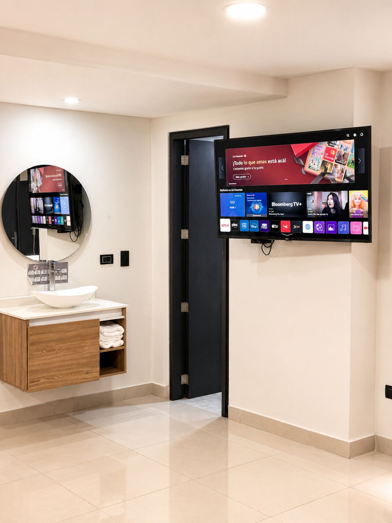 Ein modernes Badezimmer mit rundem Spiegel, Waschbecken in Weiß und einem flachbildfernseher an der Wand, der verschiedene Sender zeigt.