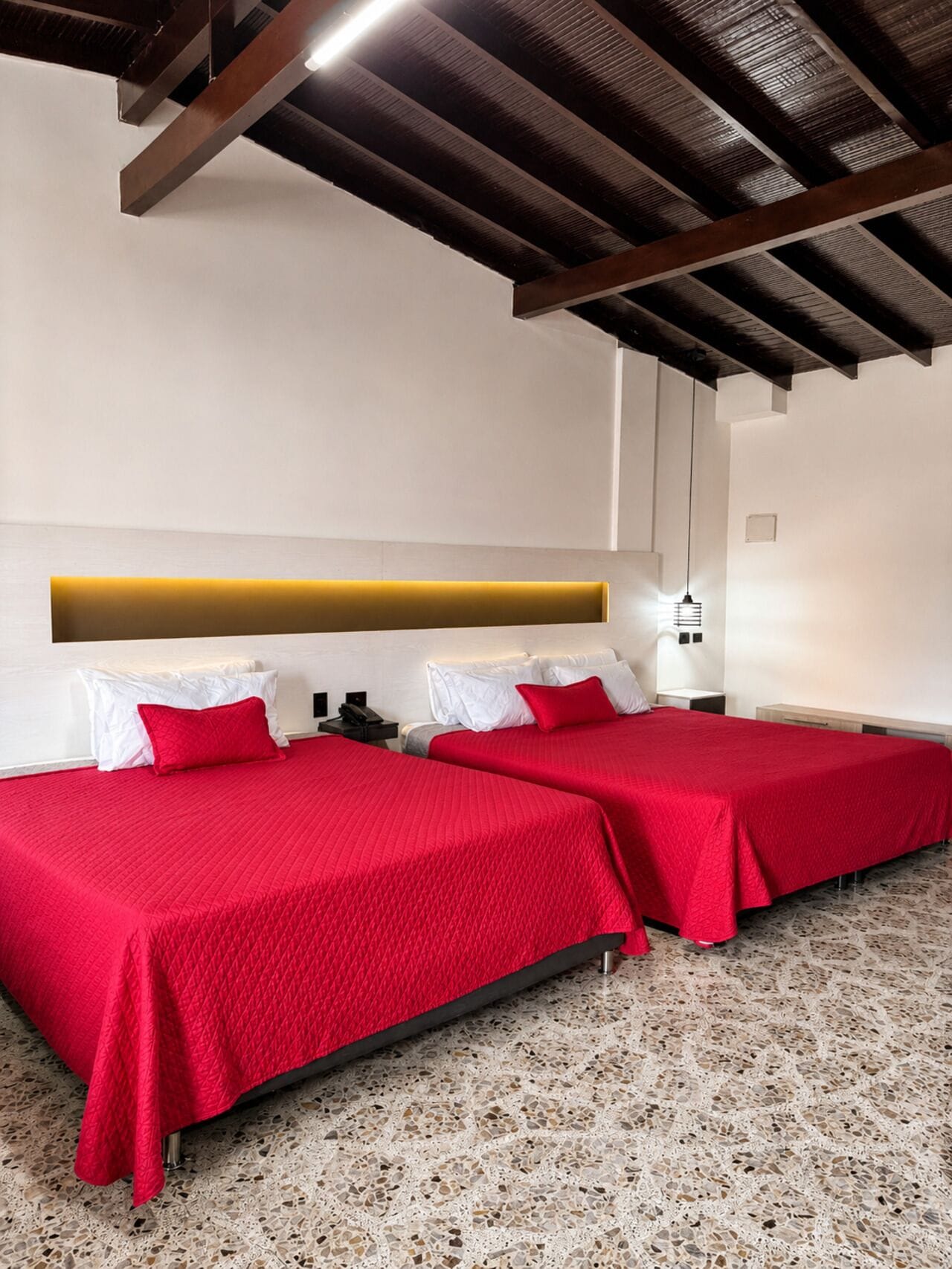 Een hotelkamer met twee bedden, witte beddengoed, een rode sierkussen, een modern hoofdbord, een klein nachtkastje en een wandlamp.
