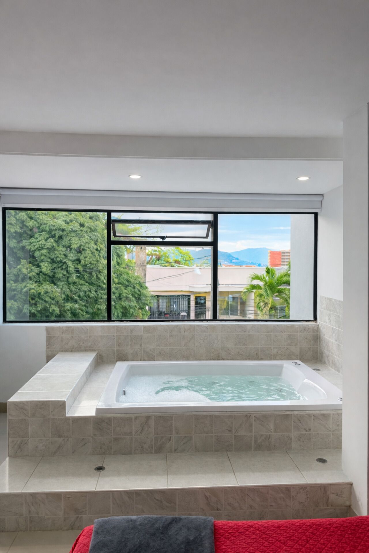Une salle de bain moderne avec une grande baignoire, une fenêtre donnant sur un paysage verdoyant et des bâtiments, et une serviette rouge sur le sol.