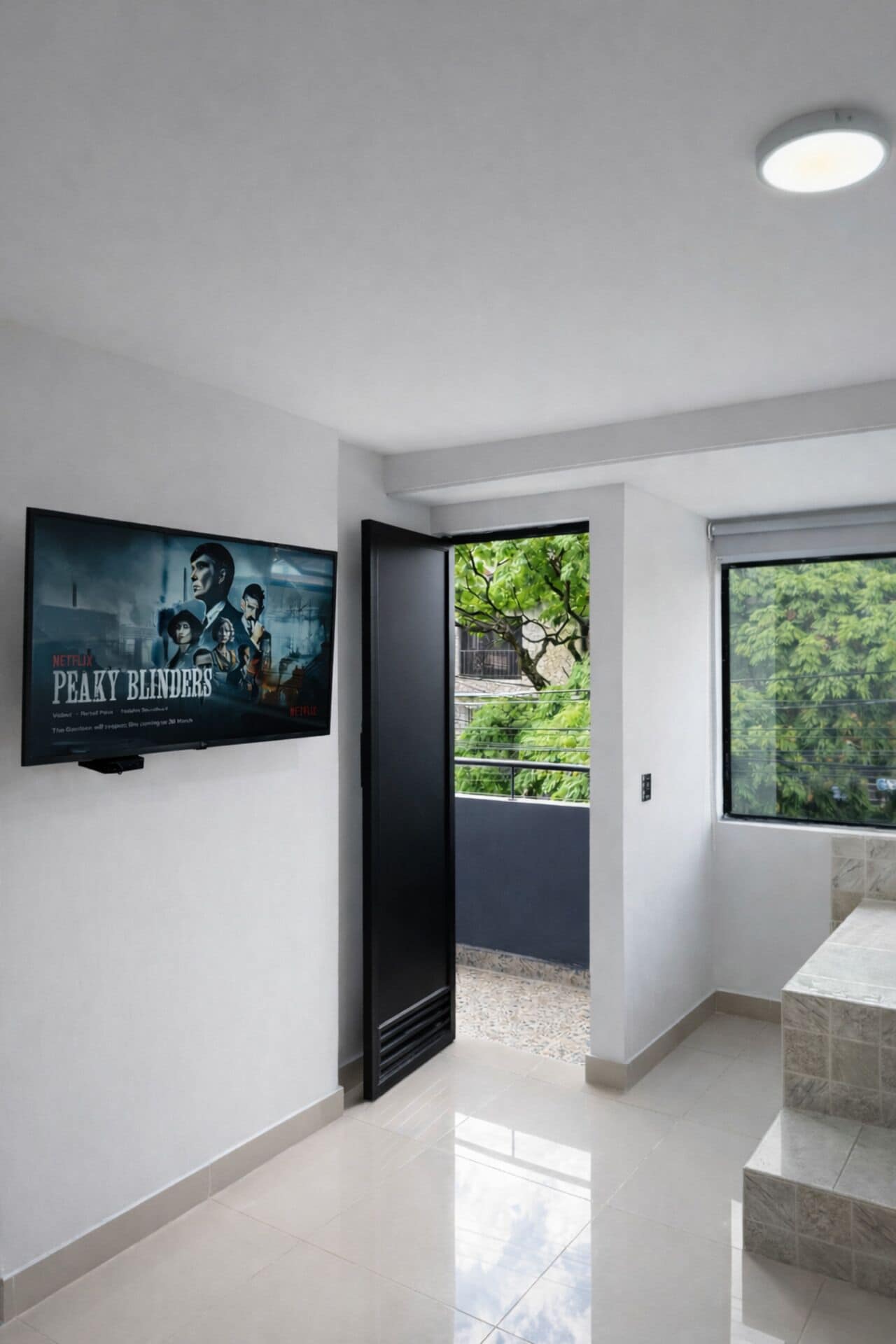 A modern interior with a flat-screen TV mounted on a white wall, a door leading to a balcony with a view of greenery, and a tiled floor.
