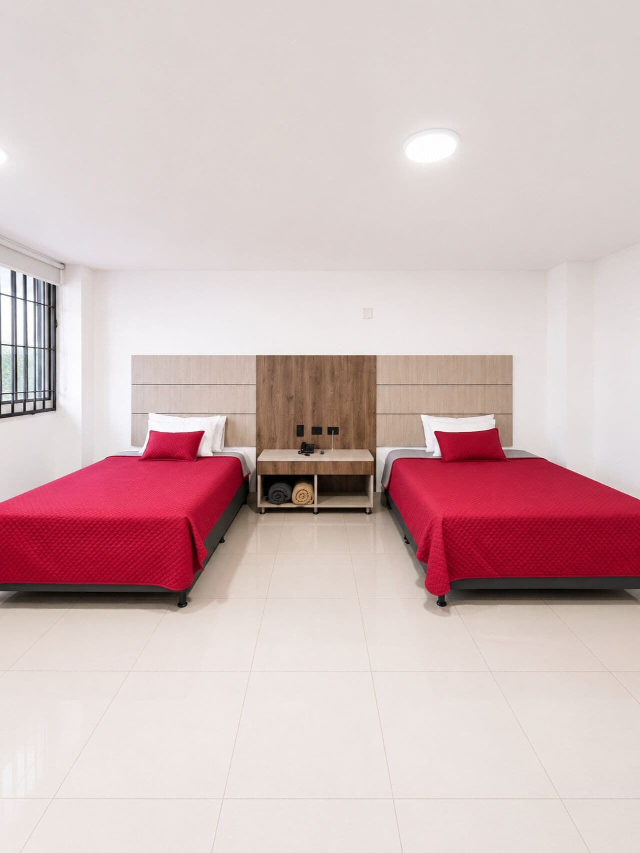 A room with two beds, a wooden headboard, a small table, and a window with bars.