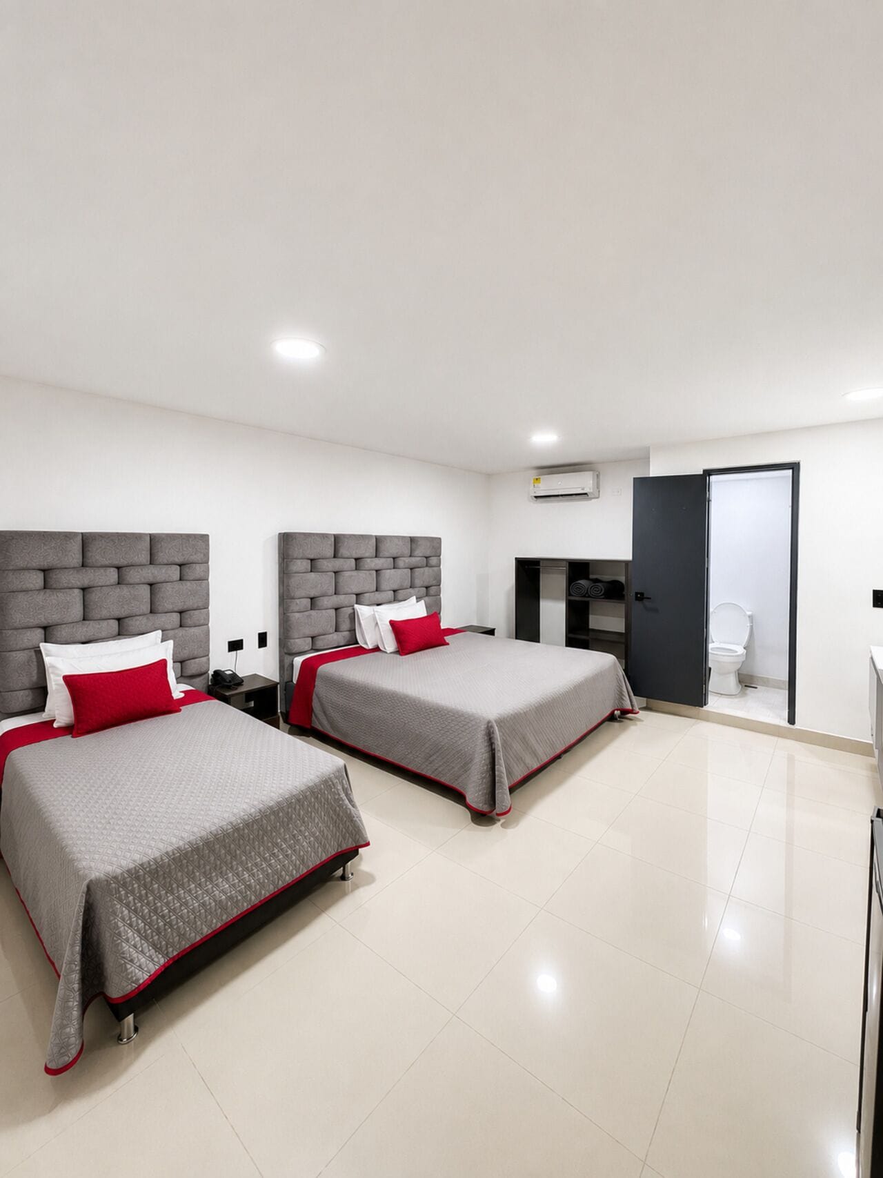 A modern hotel room with two beds, a wardrobe, a bathroom, and an air conditioning unit.