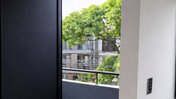 Un couloir avec une porte sombre qui donne sur un balcon offrant une vue sur un arbre et des immeubles.