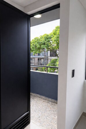 Un couloir avec une porte sombre qui donne sur un balcon offrant une vue sur un arbre et des immeubles.