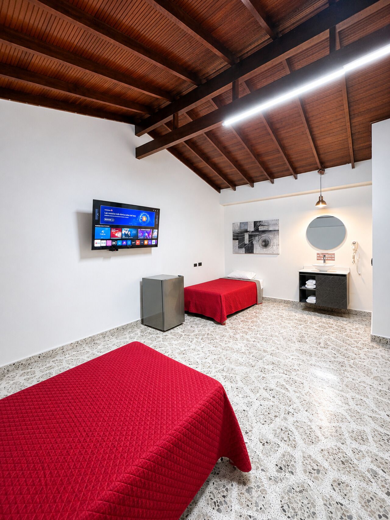 A room with two beds, a television on the wall, a small cabinet, and a ceiling with wooden beams.