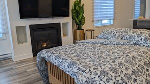 A bedroom with a large bed, a fireplace, a TV mounted on the wall, and a window with blinds.