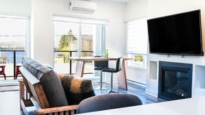 A modern living room with a sofa, a dining table with chairs, a flat-screen TV mounted on the wall, and a fireplace.