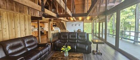 A spacious living area with a high ceiling and wooden beams, featuring a leather sofa, a coffee table with a vase of flowers, and large windows offering a view of the outdoors.
