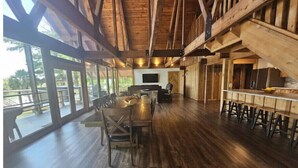Una habitació àmplia amb una taula de fusta llarga i cadires, una zona de bar amb tamborets, i grans finestres que ofereixen vista a l'exterior.